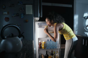 African american woman doing midnight snack at home. She eats a sandwich and looks for food into the refrigerator at night.