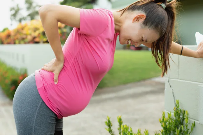 A pregnant mom with a big belly stopping to rest from exercising, bending down from a painful cramp and back unjury.