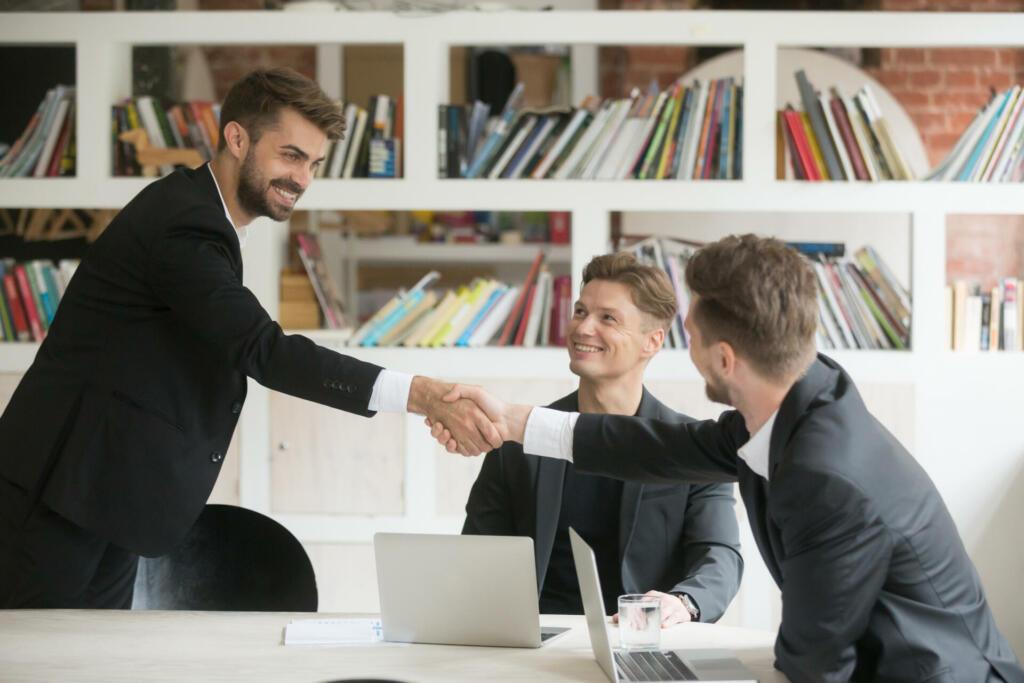 Businessman greeting congratulating new smiling executive team member or making deal with handshake at conference table, friendly company ceo shaking partners hand welcoming at meeting among three