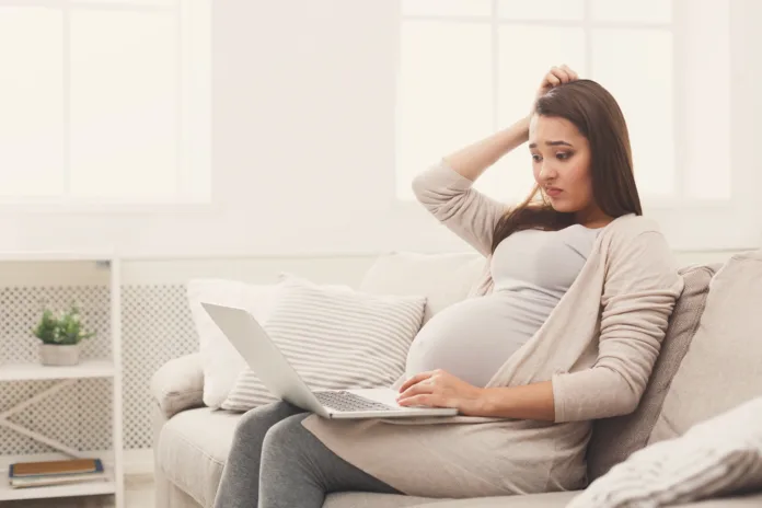 Confused pregnant woman using laptop, sitting on sofa at home. Young expectant lady surfing net, remote working, buying goods for newborn. Online shopping, pregnancy concept, copy space