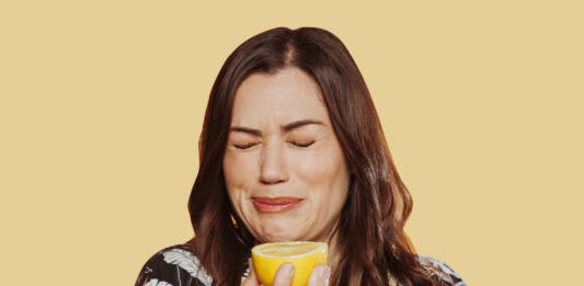 Face portrait of woman wears floral shirt, eating acid lemon doing funny face expression with eyes closed. Studio shot over yellow background.