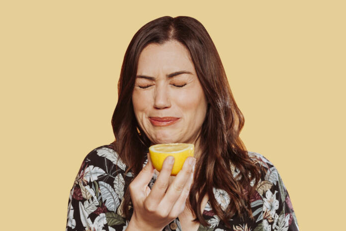 Face portrait of woman wears floral shirt, eating acid lemon doing funny face expression with eyes closed. Studio shot over yellow background.