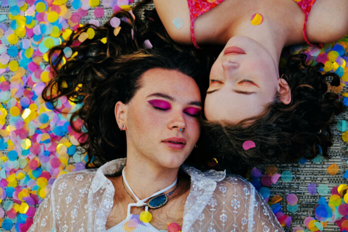 Overhead view of two non-binary friends lying down side by side, wearing bright makeup with fallen confetti and eyes closed