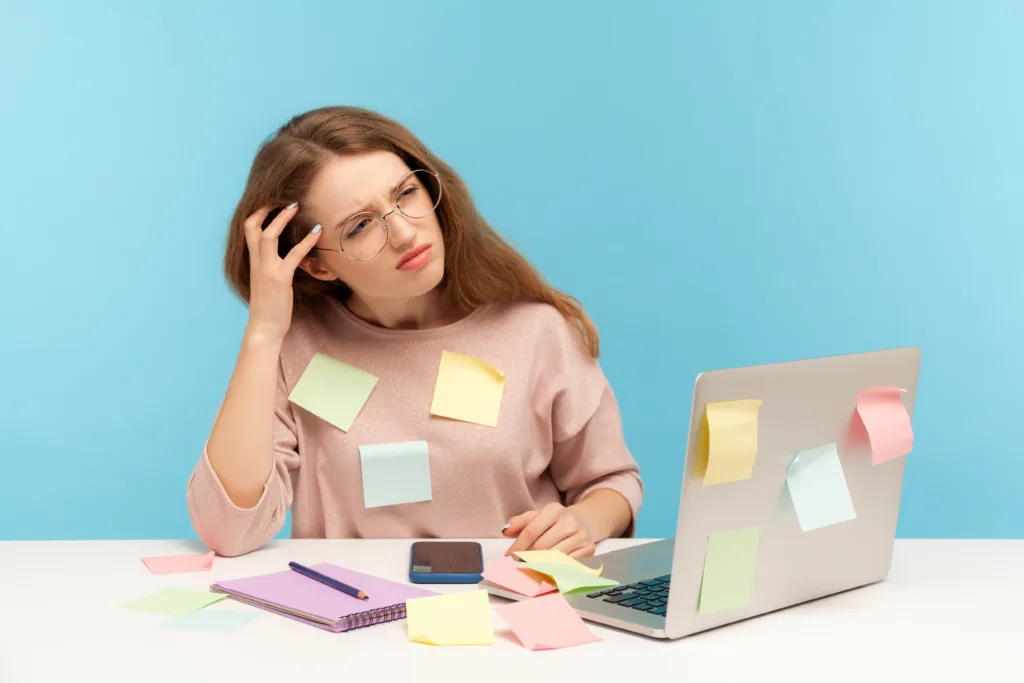 Pensive upset woman employee in nerd eyeglasses sitting at workplace office, all covered with sticky notes and thinking intensely, frustrated by workload. indoor studio shot isolated blue background