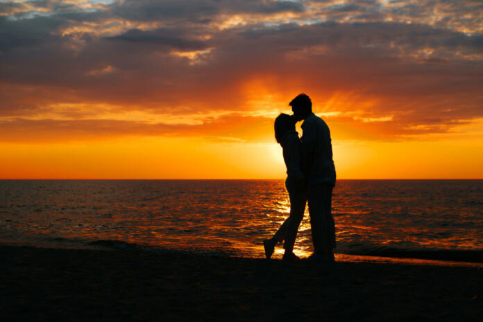 Silhouette of a happy young couple in love kissing on seashore at sunset, copy space. Flirt, romance, relationship, date, valentines day concept.