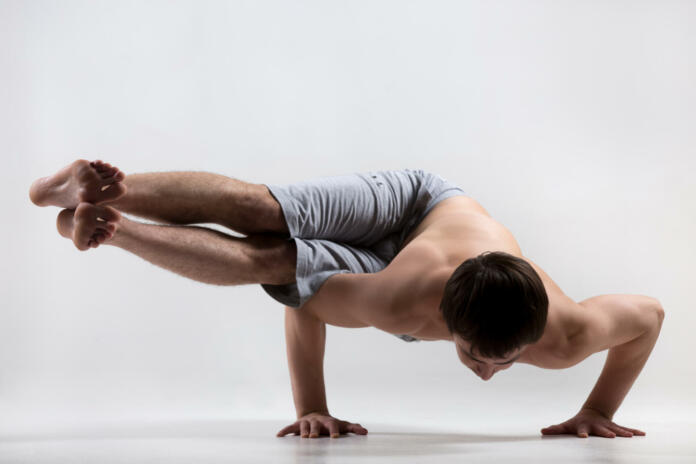 Sporty muscular young man working out, yoga, pilates, fitness training, asana Dwi Pada Koundinyasana, two-legged variation of Sage Koundinya Pose, gray background, low key shot