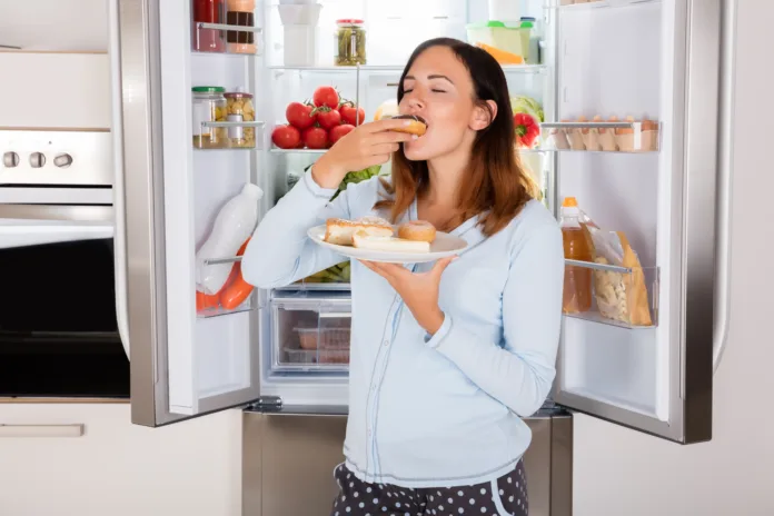 Young Woman Enjoy Eating Donut From Plate Near Refrigerator In Kitchen