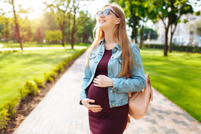 A beautiful pregnant girl student, in sunglasses, happily looks at the sky, walks in the park after studying.