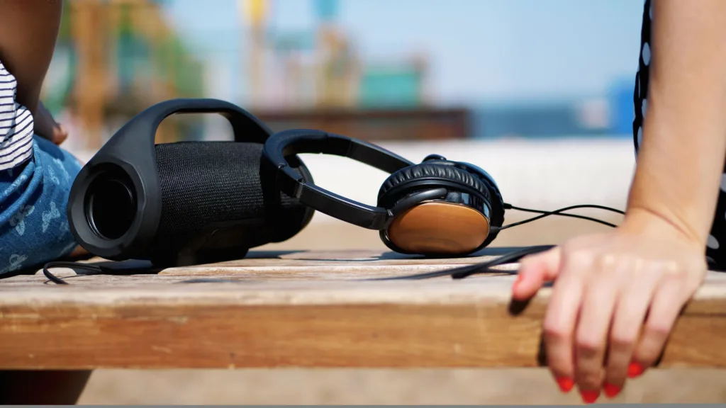 close-up, there are big headphones and a small , mini music bluetooth portable black cylinder wireless loudspeaker on the bench, in summer on the beach. High quality photo