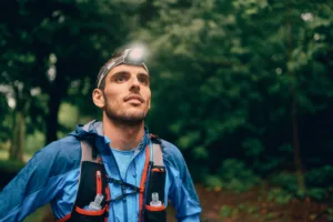 Competitive, athletic young man with a head mounted flashlight runs off road outdoors through the woods on a trail in the afternoon wearing sportswear.
