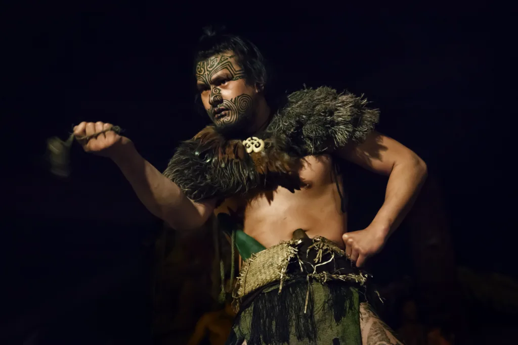 Horizontal close-up shot of a tattooed Maori warrior in traditional dress with his ancestral weapons at night, Mitai Maori Village, Rotorua, North Island, New Zealand