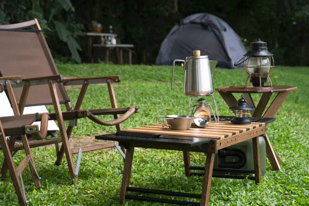 Stainless steel kettle, portable gas stove, bowl and vintage lanterns with outdoors table set on green lawn in camping area