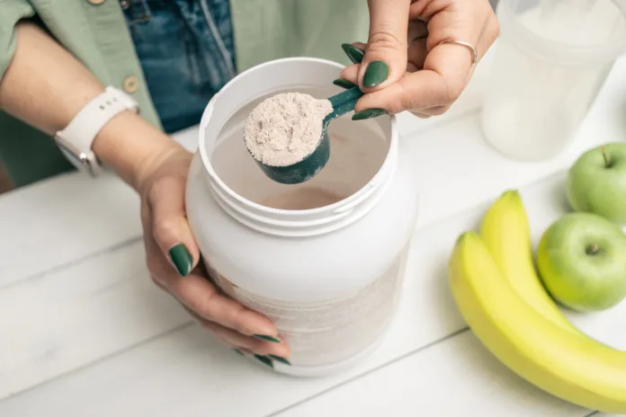 Woman in jeans and shirt holding measuring spoon with portion whey protein powder above plastic jar on white wooden table with shaker, banana and apple fruit. Process of making protein drink.