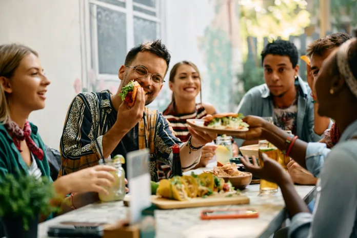 Young happy people having lunch together. Focus is on man passing tacos to his female friend.