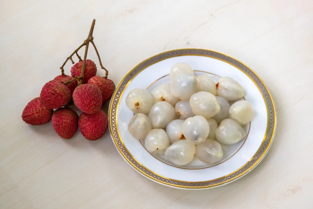 A bunch of ripe lychee and peeled litchi on a white plate on wooden surface.