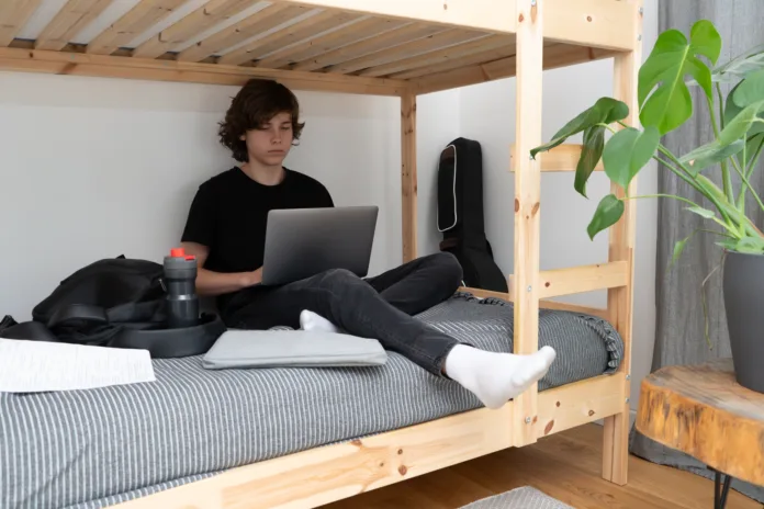 A teenage schoolboy or student is sitting on the lower tier of a bunk bed in his room with a laptop in his hands and doing homework.