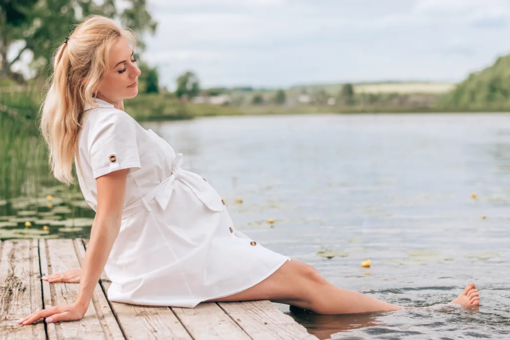 A young blonde pregnant woman in a white dress is sitting on a bridge by the river, with her feet in the water.Summer,pregnancy and prenatal care concept.