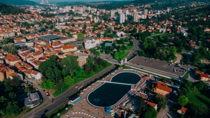 An aerial shot of the cityscape of Tuzla, Bosnia, with white buildings with brown roofs, trees, and a swimming pool