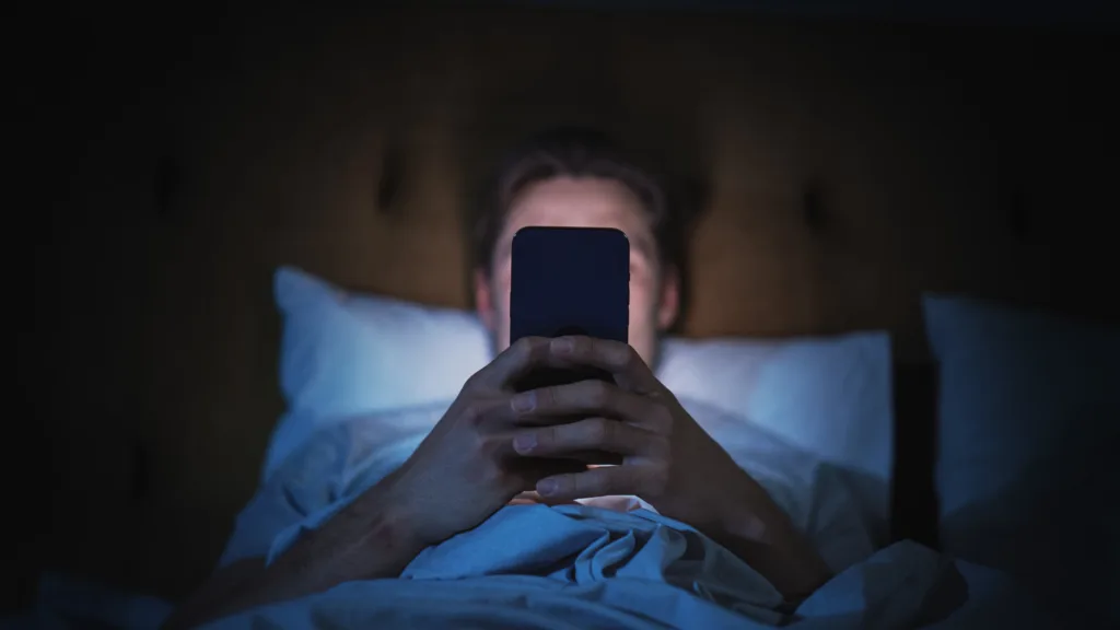 Anonymous Man Uses Smartphone in Bed at Home at Night. Handsome Guy Browsing Social Media, Reading News, Doing Online Shopping Late at Night. Focus on Hand Holding Mobile Phone Covering Face