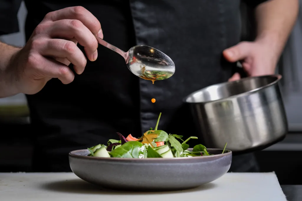 Close-up of the hands of a male chef on a black background. Pour sauce from the spoon on the salad dish. Food decoration.