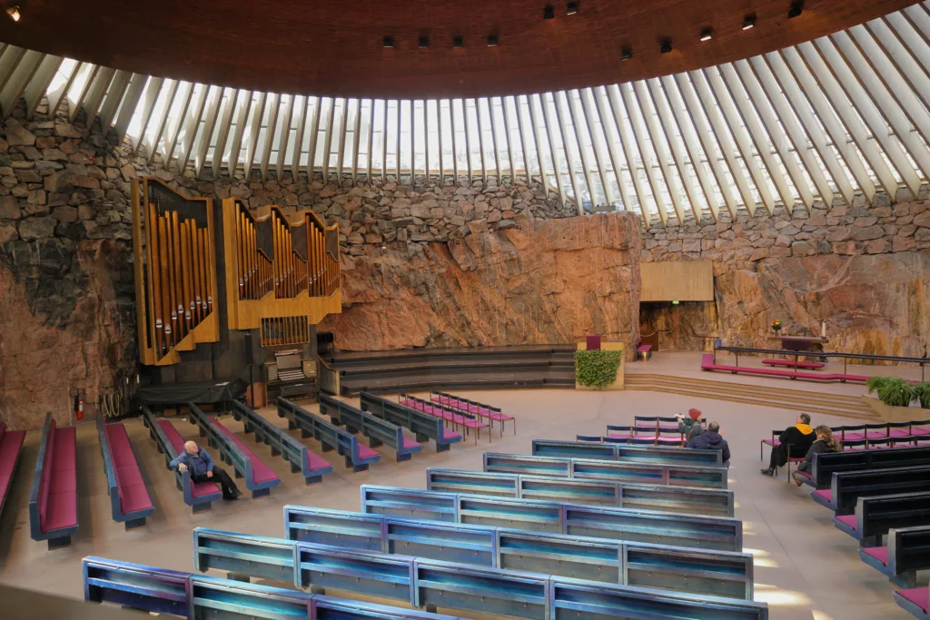 Interior of Temppeliaukio Church with seating, pipe organ, altar and copper roof in view. The church is also known as the Church of the Rock and is located in the Töölö neighbourhood of Helsinki, Finland. The church, designed by architects and brothers Timo and Tuomo Suomalainen, opened in 1969 and is built directly into solid rock