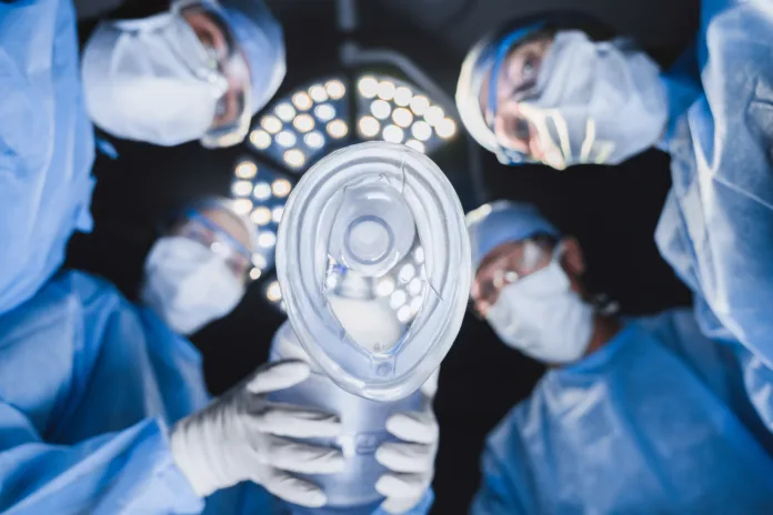 Low angle view of anesthetist holding oxygen mask above patient in surgery room with team of surgeon professionals.