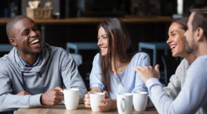 Multiracial friends girls and guys having fun laughing drinking coffee tea in coffeehouse, happy diverse young people talking joking sitting together at cafe table, multicultural friendship concept