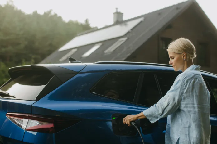 Portrait of woman charging electric car in front of her house with solar panels on roof. Mother plugging charger into the charging port. Solar energy and sustainable lifestyle of young family.