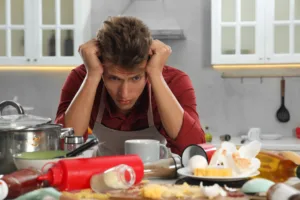 Upset man in messy kitchen. Many dirty dishware, utensils and food leftovers on table