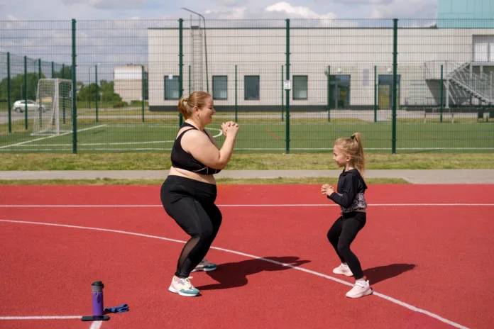 Young mother and skinny daughter play sports together, doing squat exercises on sports field. Plump woman with merrily laughing child. Girl supports mother in effort to lose weight. Outdoor fitness