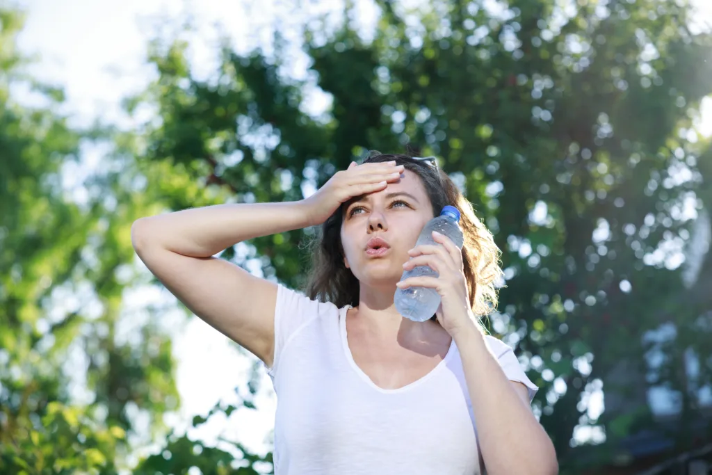 Young woman having hot flash and sweating in a warm summer day. Woman touching face with cold water bottle to cool off