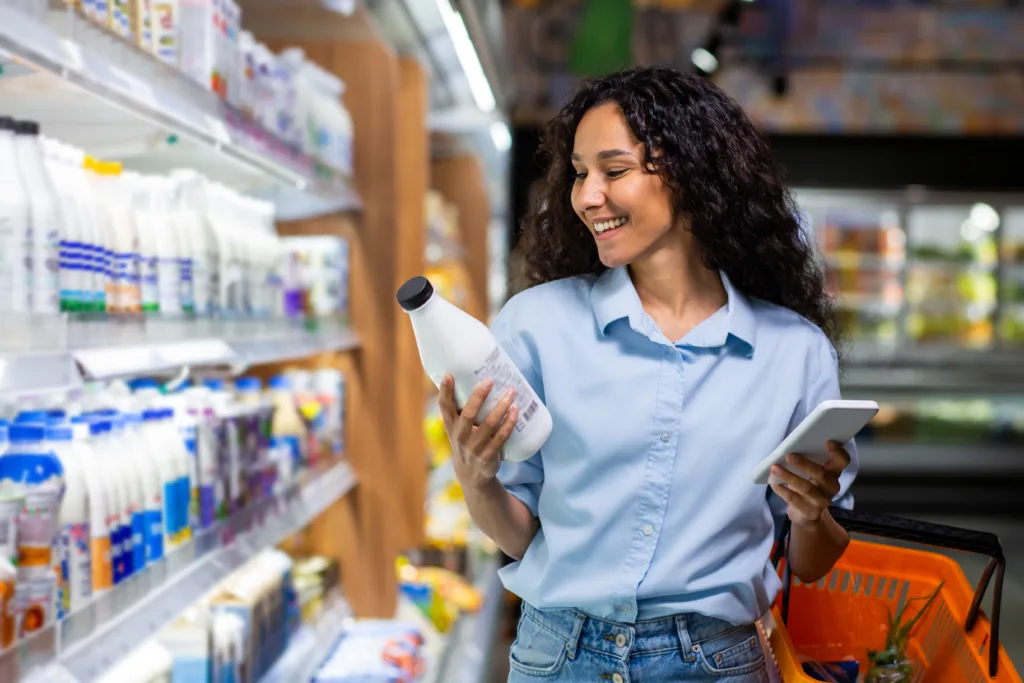 Cheerful young woman with curly hair holding a product and a digital tablet while shopping in a grocery store aisle.