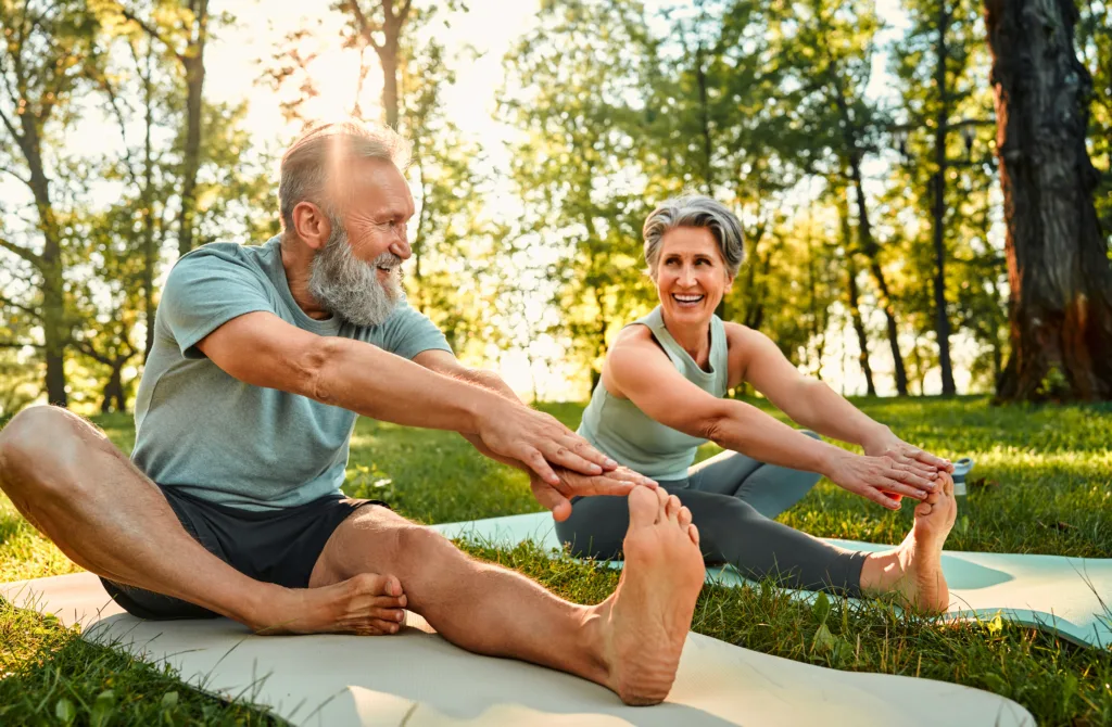 Flexible exercises for body. Sporty man and woman with grey hair stretching on yoga mats with hands to one leg during outdoors workout. Happy married couple with bare feet warming up together at park.