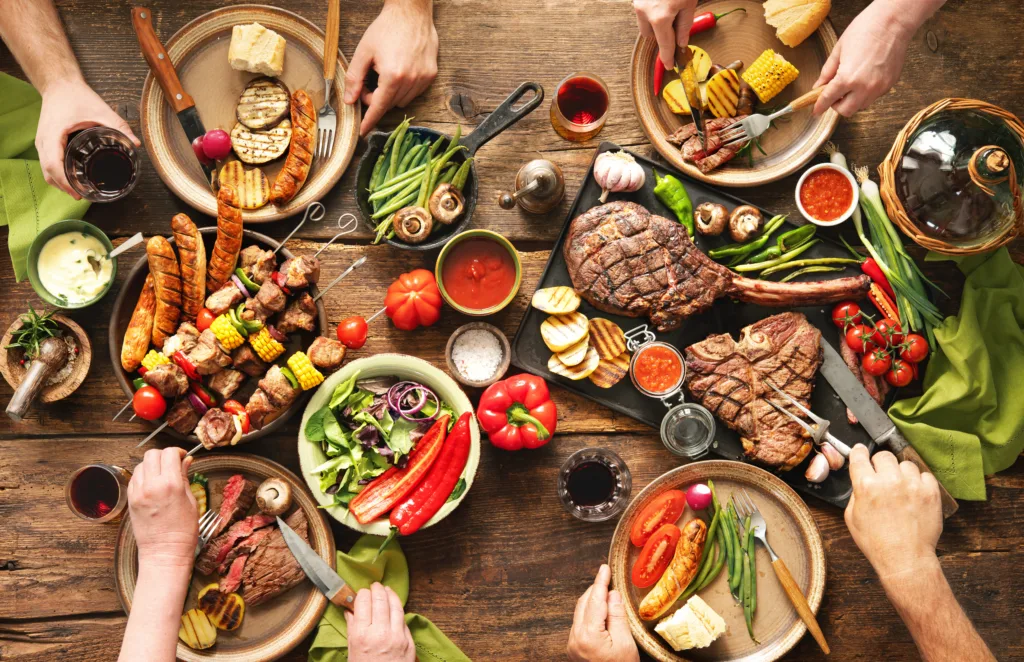 Friends having a barbecue party. Group of people eating grilled meats and vegetables and drinking wine at rustic picnic table