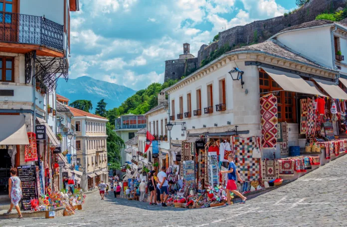 Gjirokaster town, Albania –August 2, 2020: summer cityscape – street of ancient town with souvenir shops and tourists. Old historical architecture and Castle complex on the hill.