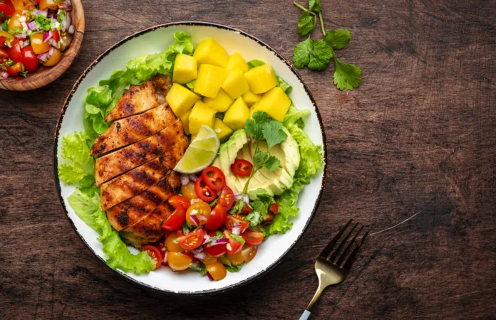 Grilled chicken with side dish salad from avocado, mango, tomato salsa, cilantro, lime and lettuce, rustic wooden table background, top view