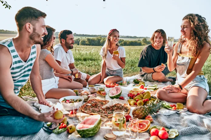 Group of young attractive friends having a picnic, sitting on a grey blanket on green grass