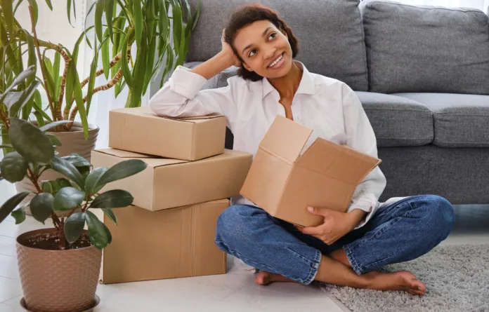 Happy young woman sitting in new apartment and raising arms in joy after moving in. Joyful and excited african girl moving to new home. Black woman sitting on floor in her house.