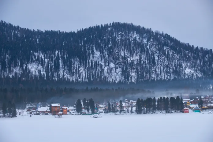 Houses on the bank of frozen Teletskoe lake under the chimney smoke. Iogach, Altai, Siberia, Russia