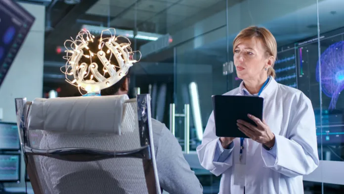 Man Wearing Brainwave Scanning Headset Sits in a Chair while Scientist with Tablet Computer Supervises Process. In the Modern Brain Study Laboratory Monitors Show EEG Reading and Brain Model.