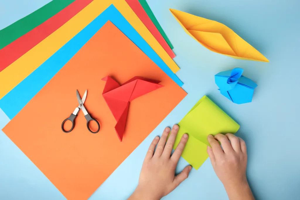 Origami art. Child folding paper on light blue background, closeup and top view