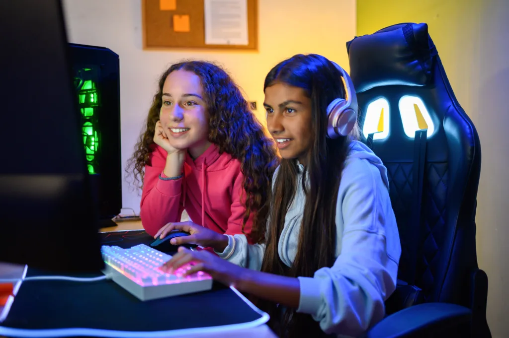 Positive teenage multiracial looking at screen of monitor while sitting at table with gaming PC and using keyboard to play game in room