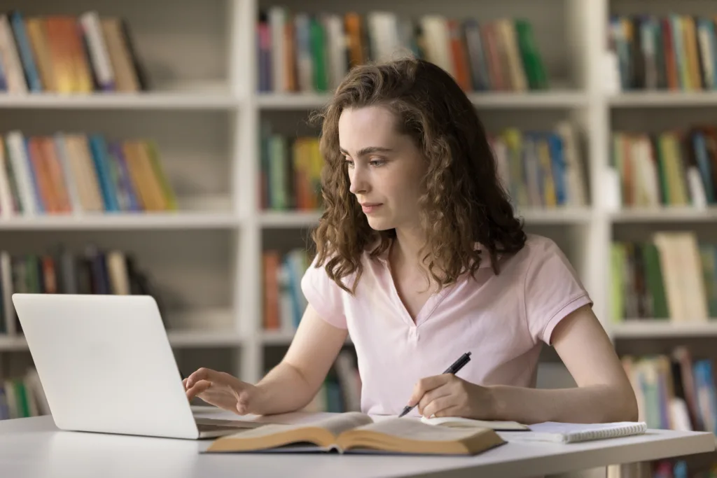 Serious female student studying on-line, take notes, writing essay sit at desk with laptop and textbook. Generation Z using internet resources, modern technology for e-learning, gaining new knowledge