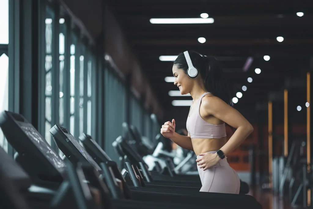 Side view of beautiful young asian woman running on treadmill and listening to music via headphone during sports training in a gym.