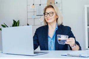 Smiling mature businesswoman working on laptop and drinking coffee at office, confident middle-aged experienced senior female professional, CEO of company. Female entrepreneur manage business