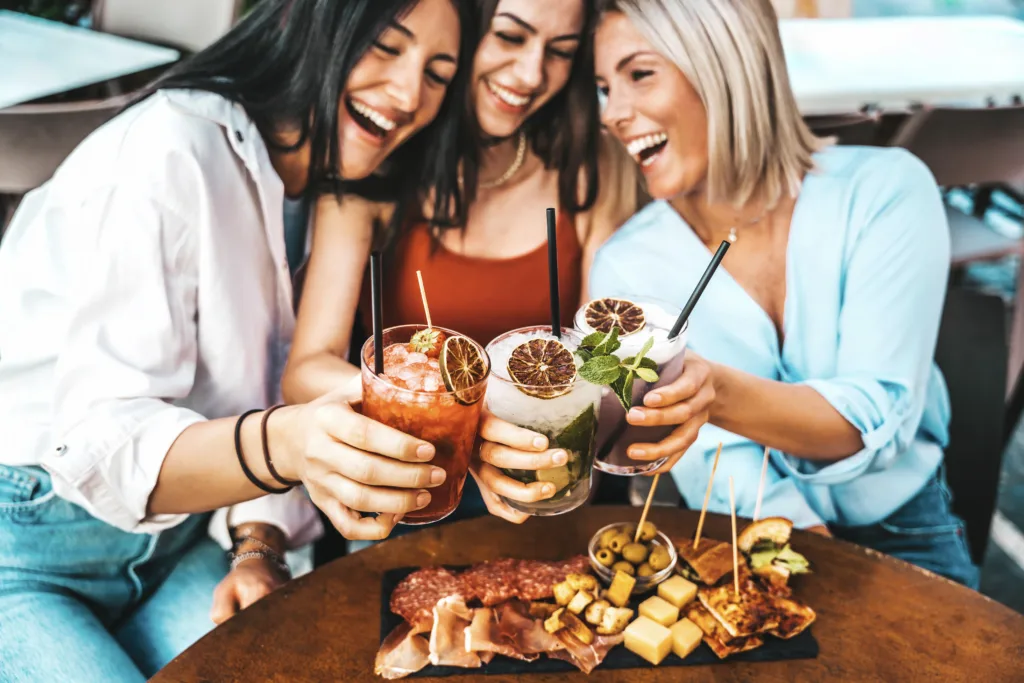 Three beautiful young women drinking cocktails sitting at bar restaurant table - Happy female friends enjoying summertime holidays outdoors - Life style concept with girlfriends hanging out together