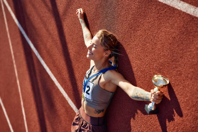 Winner woman in sportswear with number with gold medal for first place and cup lying at stadium celebrating after competition. Sports and  victory concept.