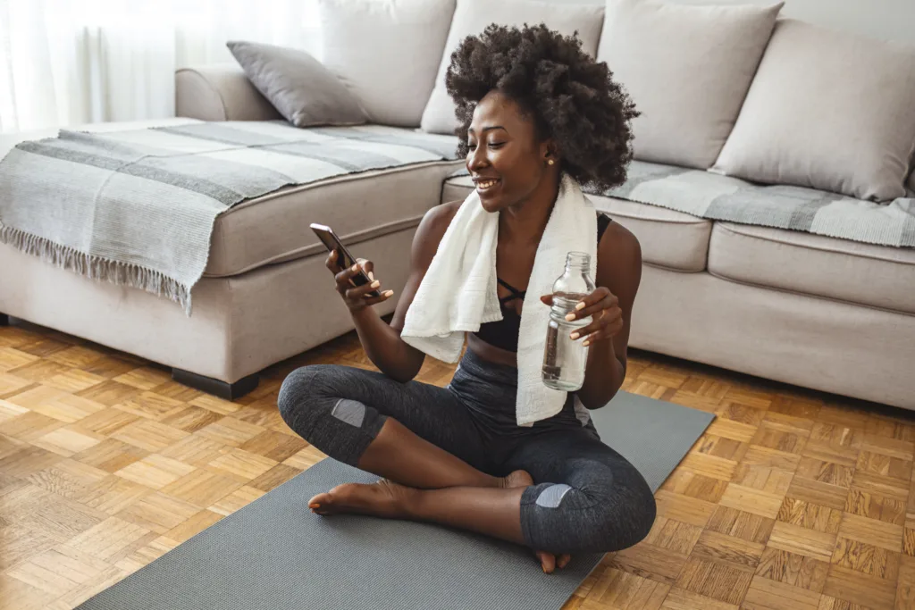 Woman using smart phone on exercise mat in front of her laptop. Sporty girl is exercising at phone on mat enjoying playlist on mobile phone. Positive active energtic sportswoman sit on purple mat use cellphone