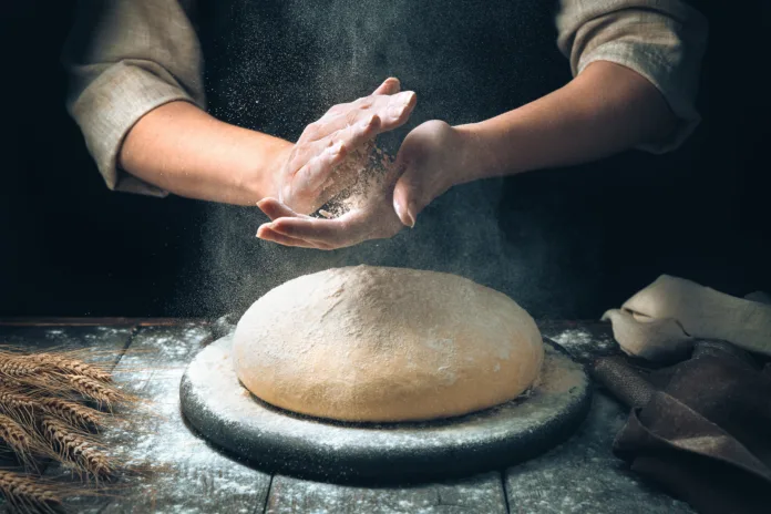 Women's hands knead the dough, from which they will then make bread, or pizza. A cloud of flour flies around like dust.