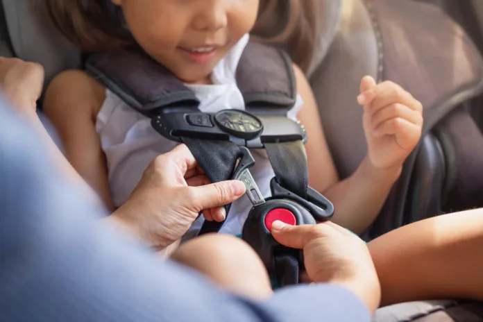 A toddler sitting in the child car seat with the mother helping to buckle and fasten seat beat properly to stay safe while driving.